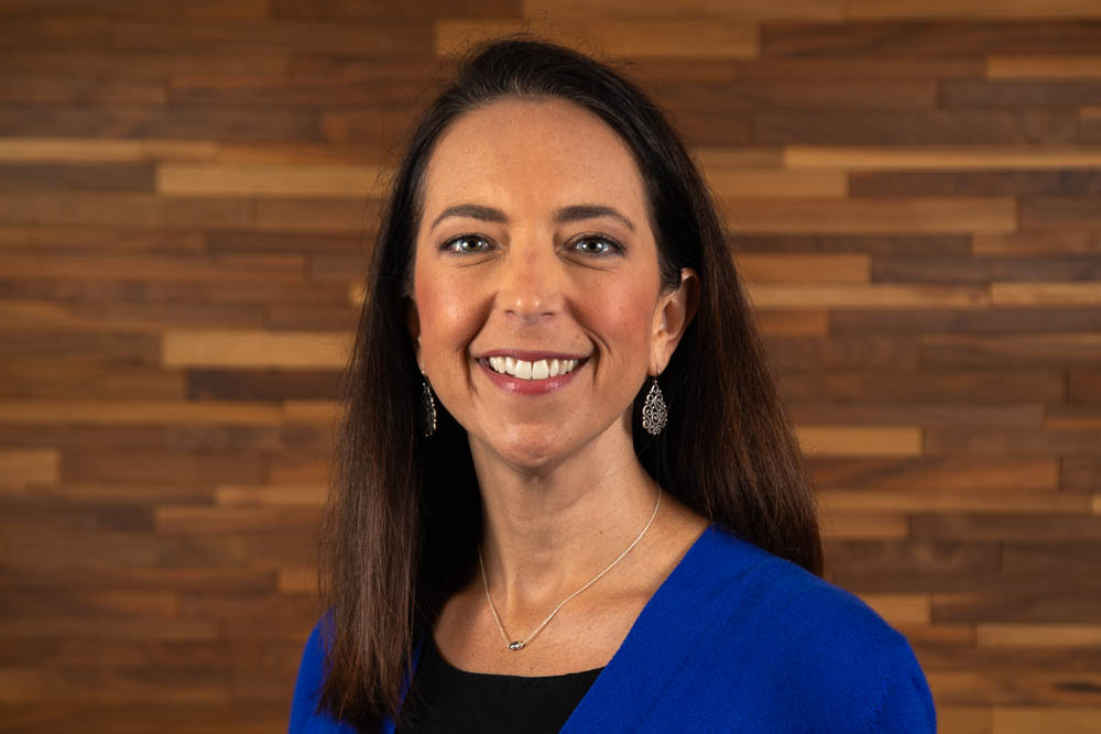 Headshot of Meredith Simpson dressed in a blue dress shirt in front of a wooden background.