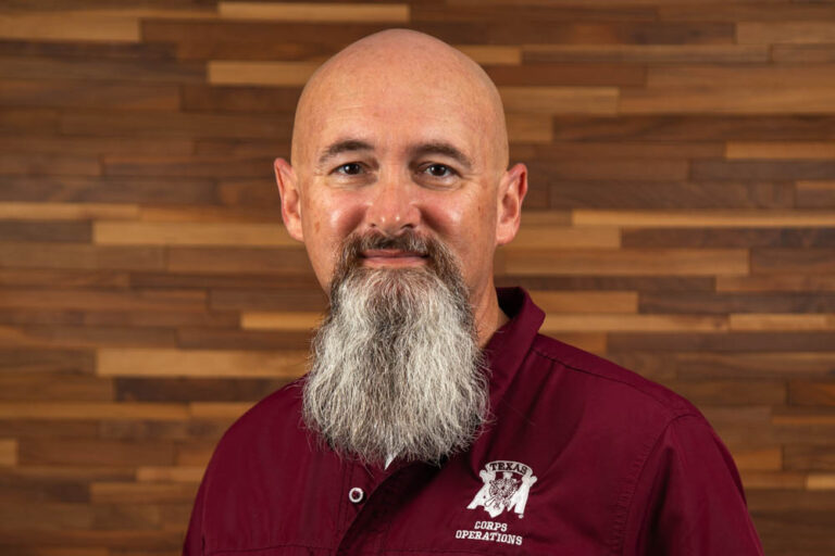 Headshot of John Reagan dressed in a maroon collared shirt in front of a wooden background.