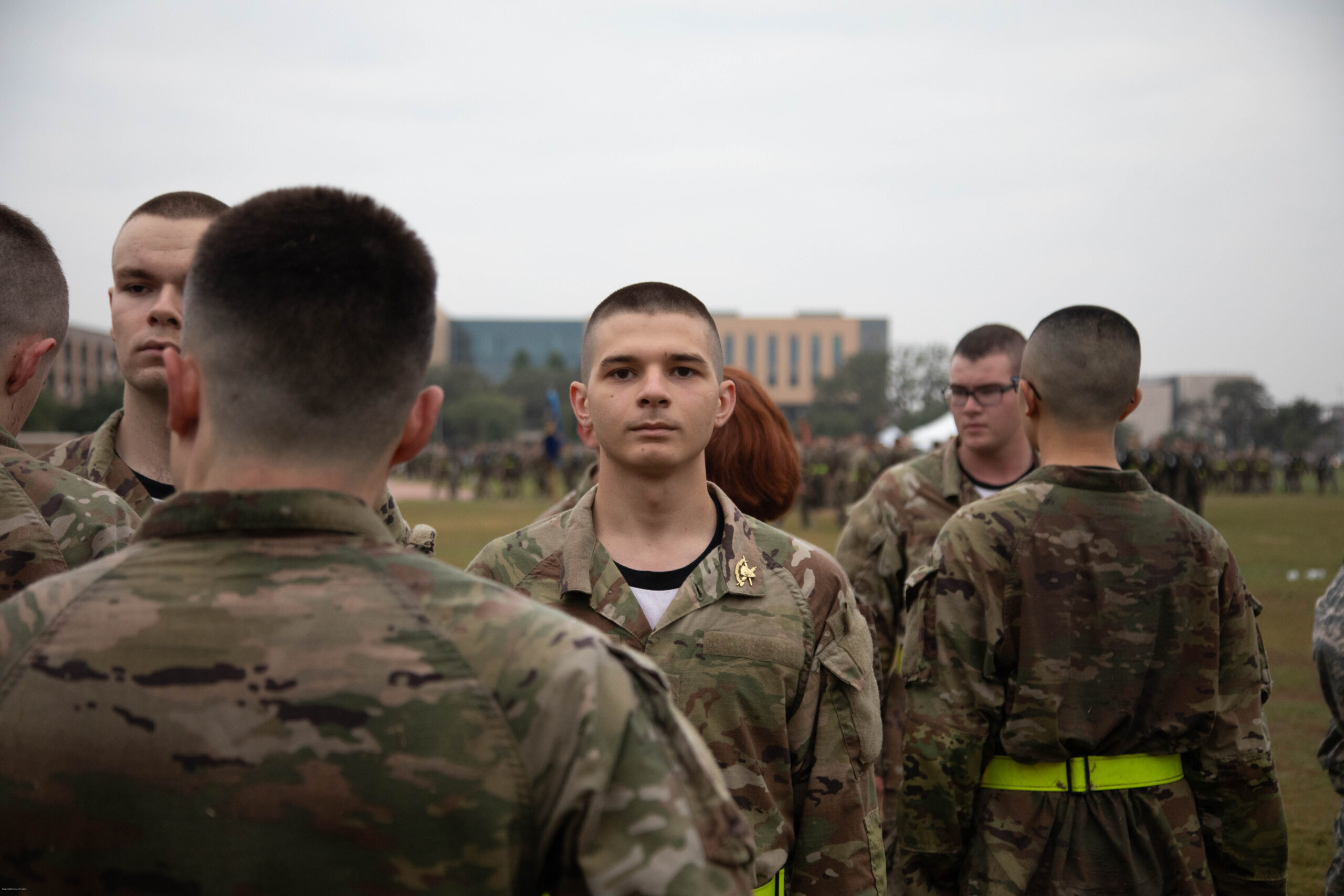 A group of soldiers in camouflage uniforms stand in formation outdoors, with one young male soldier facing the camera directly. Buildings and more people are visible in the blurred background.