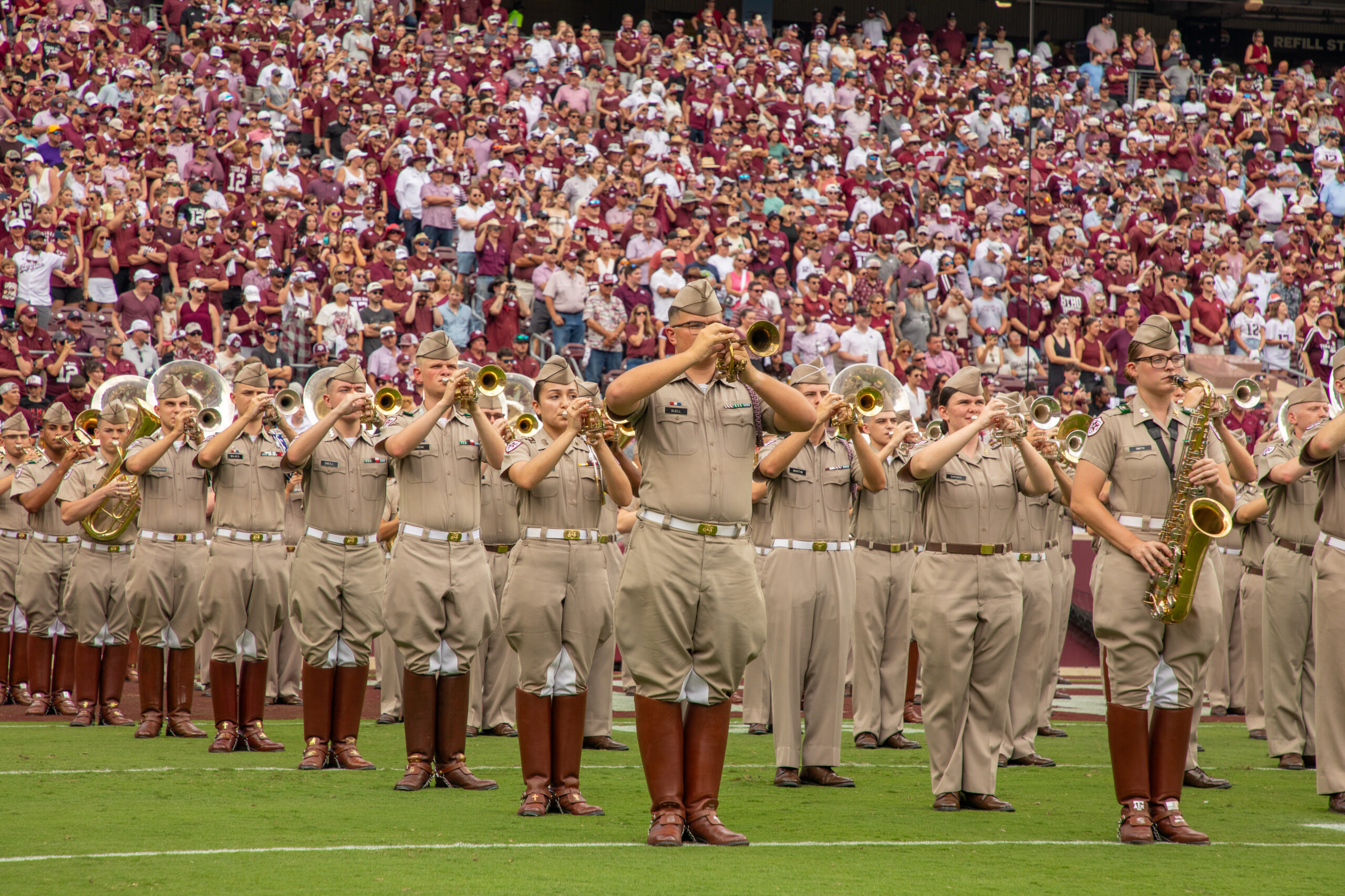 A marching band in tan uniforms and brown boots performs on a football field, playing brass and woodwind instruments, with a large crowd of fans in maroon and white filling the stadium bleachers behind them.