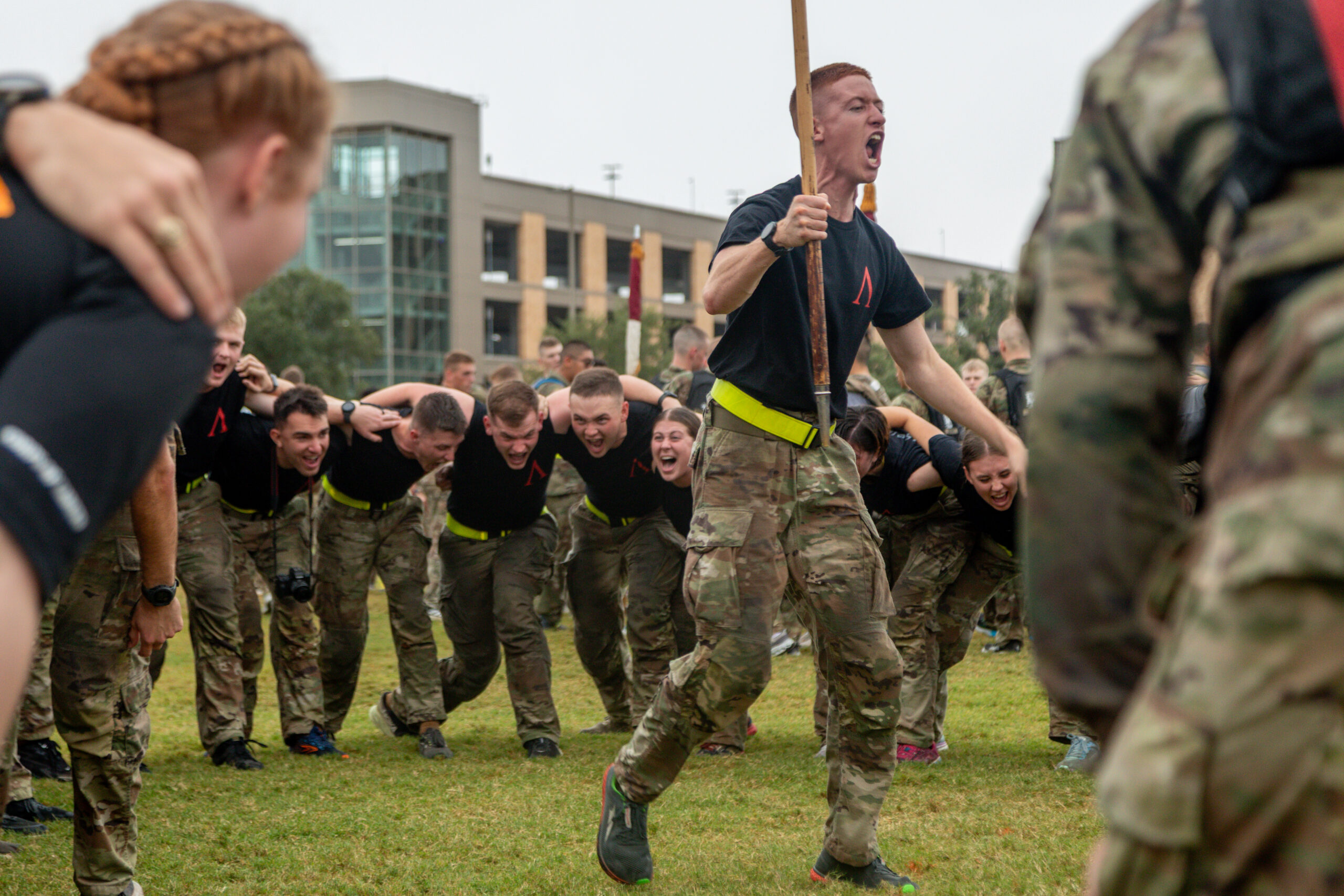 A group of soldiers in camouflage pants and black shirts participate in a team-building exercise outdoors, bending forward and linked together, while one person leads, shouting and holding a wooden stick.