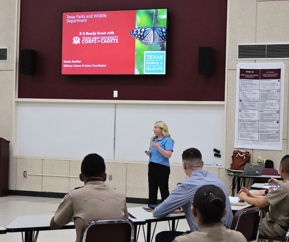 A woman stands at the front of a classroom giving a presentation to students in uniform. A large screen behind her displays a butterfly image and the Texas Parks and Wildlife Department logo.