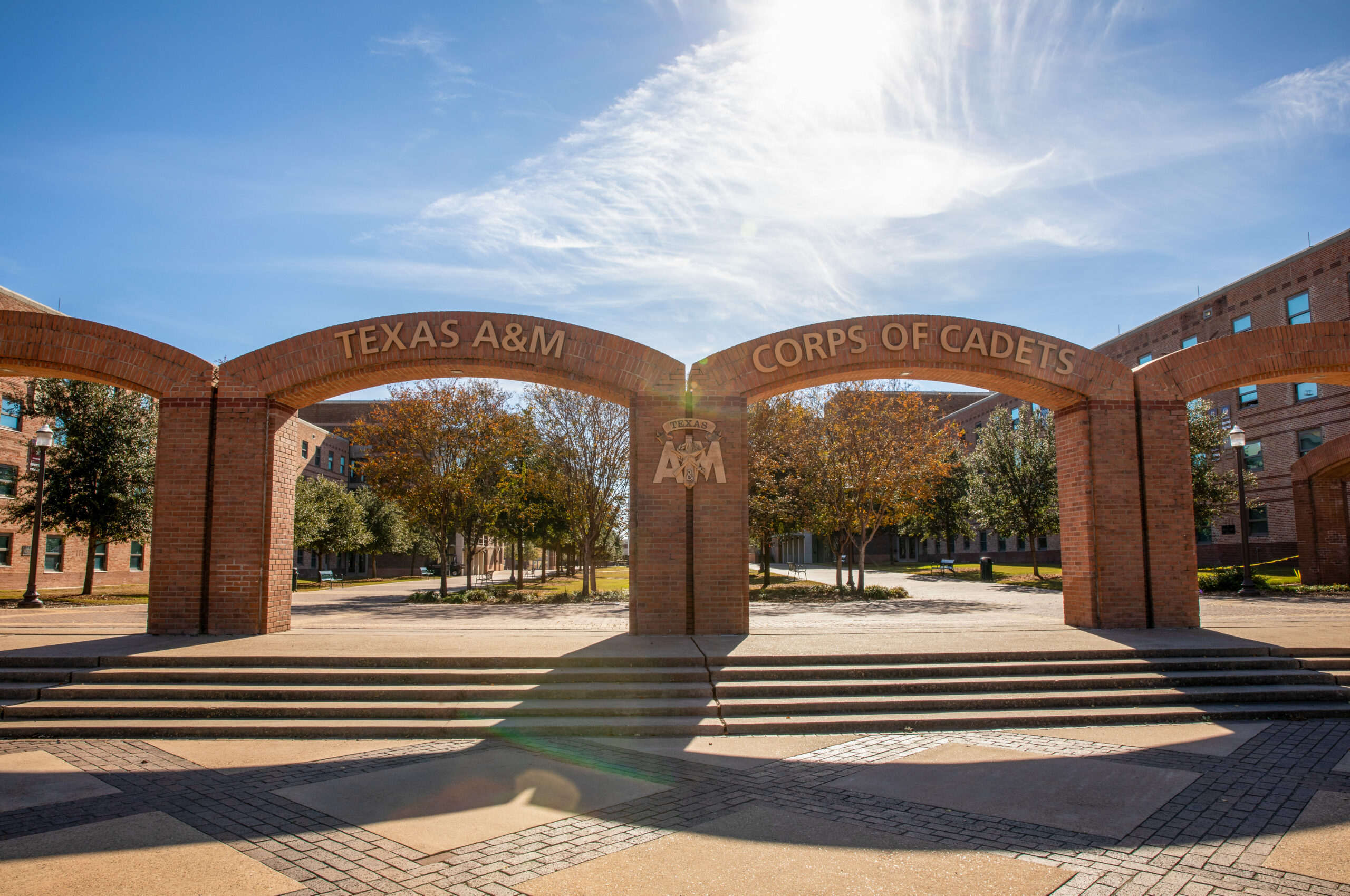 Three brick arches labeled Texas A&M and Corps of Cadets stand at an entrance to a campus plaza, with sunlight streaming through trees and buildings in the background.
