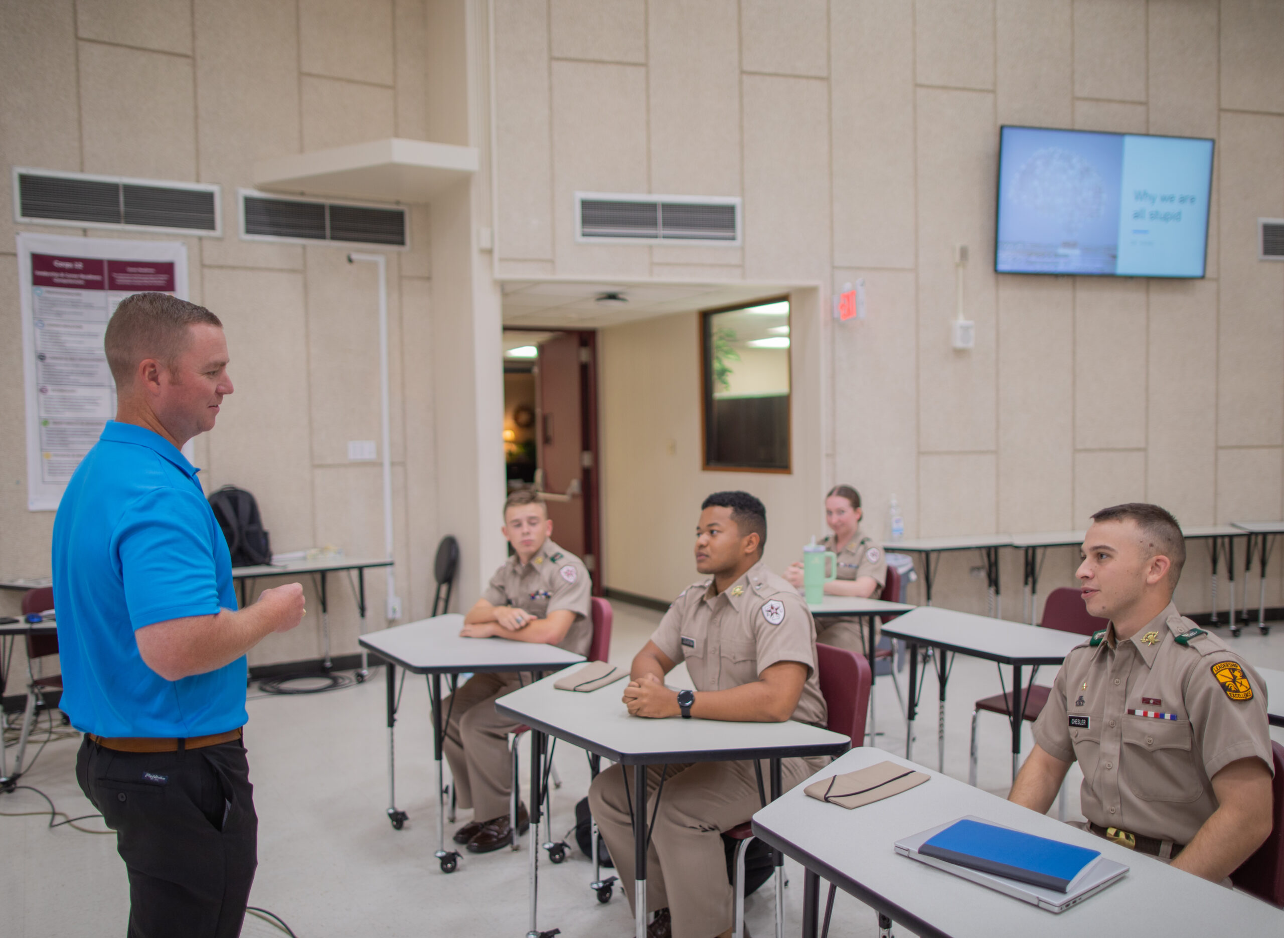 A man in a blue shirt speaks to three uniformed cadets seated at desks in a classroom. The cadets are listening attentively, and notebooks are on the desks. A large screen and posters are visible in the background.