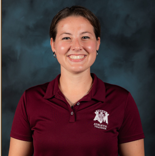 A woman with short brown hair, wearing a maroon polo shirt with an Athletic Trainer logo, smiles in front of a dark, cloudy studio background.