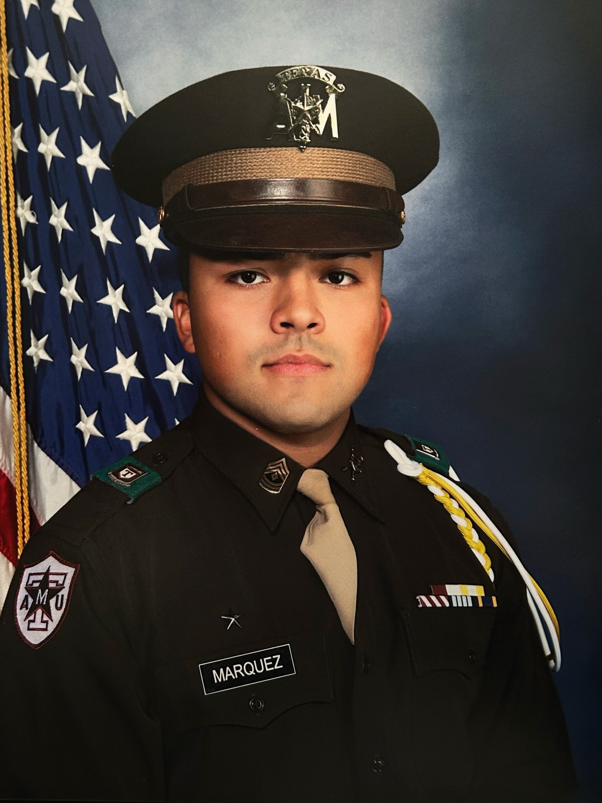 Cadet Matthew Marquez '24 poses in uniform in front of the U.S. flag and a blue, studio backdrop